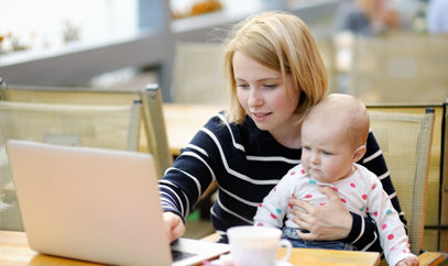 Junge Frau mit Baby im Arm vor einem Laptop